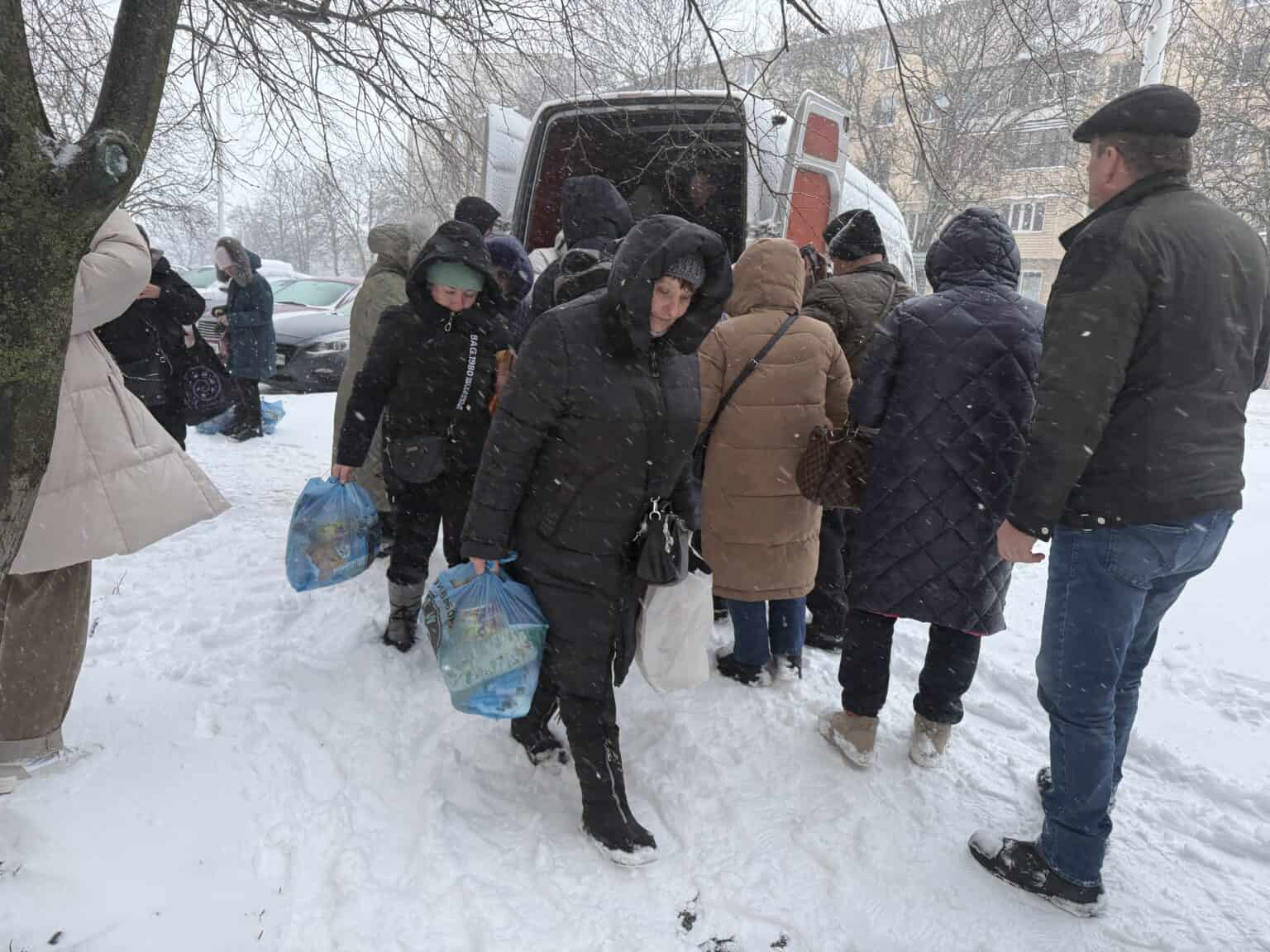 Eine Gruppe von Menschen in Wintermänteln versammelt sich im Schnee um einen Lieferwagen, einige tragen Taschen mit Hilfsgütern. Es schneit und der Boden ist bedeckt, im Hintergrund sind kahle Bäume und Gebäude zu sehen.