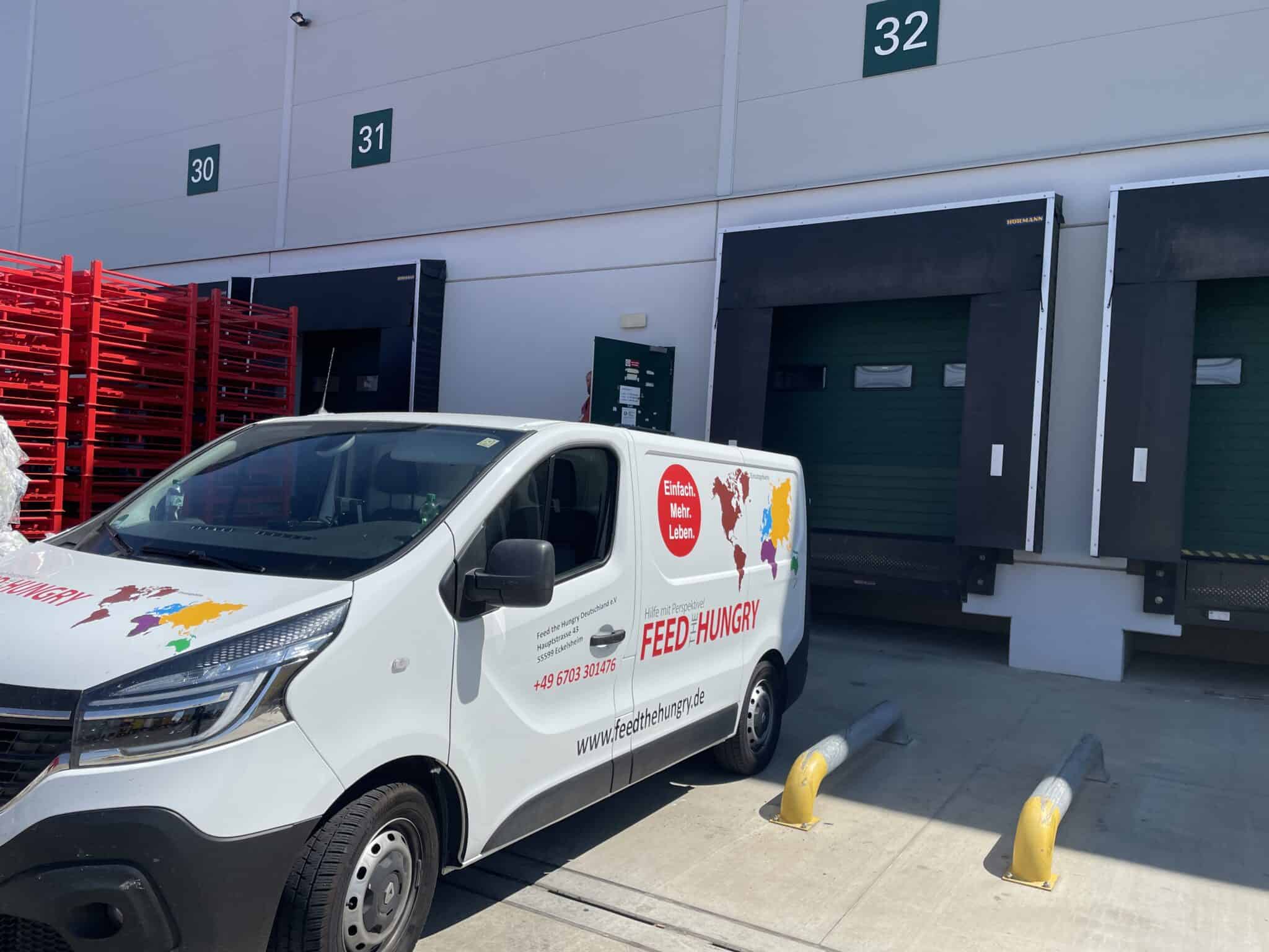 Ein weißer Transporter von FEED HUNGRY parkt vor den Laderampen mit den Nummern 30, 31 und 32 in einem Lagerhaus, in der Nähe befinden sich rote Lagerregale und das Sonnenlicht wirft Schatten auf die Szene.