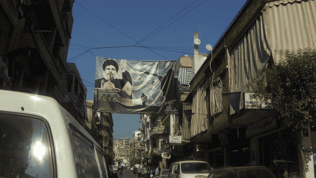 Eine Straßenszene in einer Stadt im Nahen Osten zeigt Autos, die entlang schmaler Gebäude geparkt sind, und ein großes Banner, das einen Mann in traditioneller Kleidung zeigt, hängt über der Straße. An den Gebäuden sind Satellitenschüsseln und Schilder zu sehen.