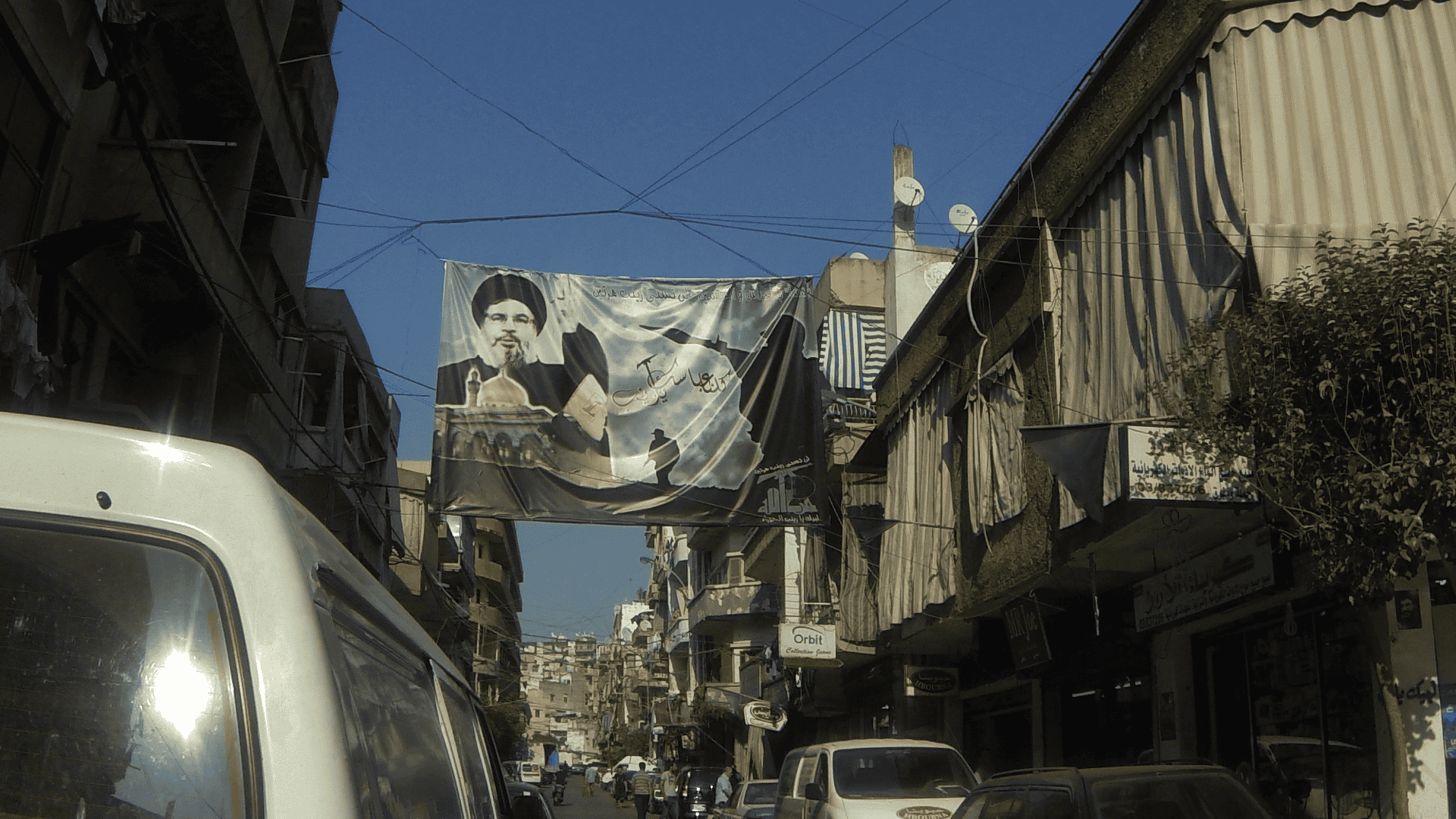 Eine Straßenszene in einer Stadt im Nahen Osten zeigt Autos, die entlang schmaler Gebäude geparkt sind, und ein großes Banner, das einen Mann in traditioneller Kleidung zeigt, hängt über der Straße. An den Gebäuden sind Satellitenschüsseln und Schilder zu sehen.