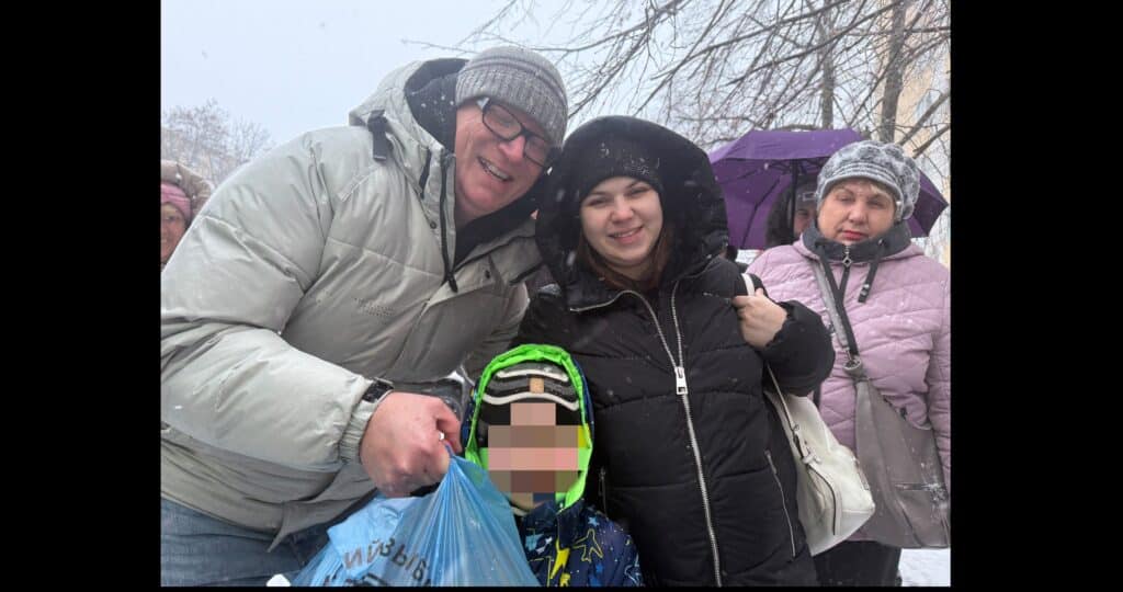 Ein Mann und eine Frau in Wintermänteln lächeln und posieren mit einem Kind mit verschwommenem Gesicht, das eine blaue Tasche hält. Um sie herum fällt Schnee, und in der Nähe stehen zwei weitere Personen in Winterkleidung. Im Hintergrund sind kahle Baumzweige zu sehen.