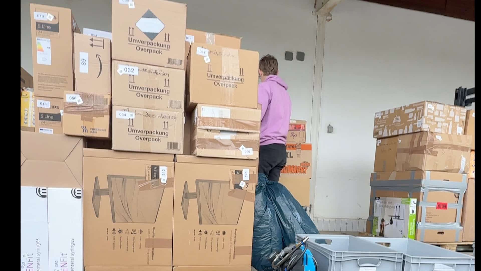 Eine Person in einem rosafarbenen Kapuzenpulli steht zwischen Stapeln von Pappkartons und Plastikbehältern, von denen einige mit "Umverpackung Overpack" beschriftet sind, in einem Bereich, bei dem es sich anscheinend um einen Lager- oder Umzugsbereich handelt.