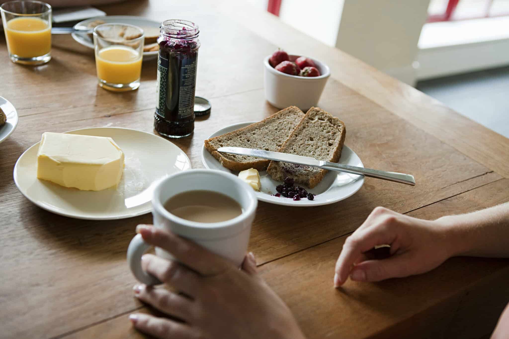 Eine Hand hält eine Tasse Tee oder Kaffee neben einem Holztisch, auf dem Vollkornbrot, Butter, Marmelade, eine Schale Erdbeeren und ein Glas Orangensaft stehen und eine gemütliche Frühstücksszene schaffen.