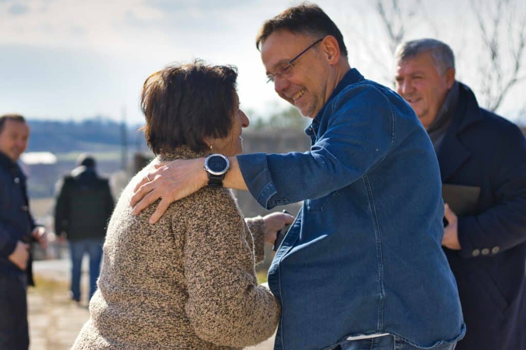 Ein Mann lächelt und umarmt im Freien eine ältere Frau, während zwei weitere Männer an einem sonnigen Tag im Hintergrund stehen.