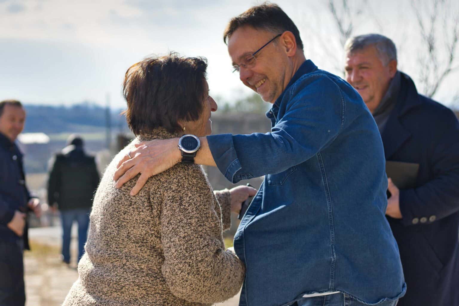 Ein Mann lächelt und umarmt im Freien eine ältere Frau, während zwei weitere Männer an einem sonnigen Tag im Hintergrund stehen.