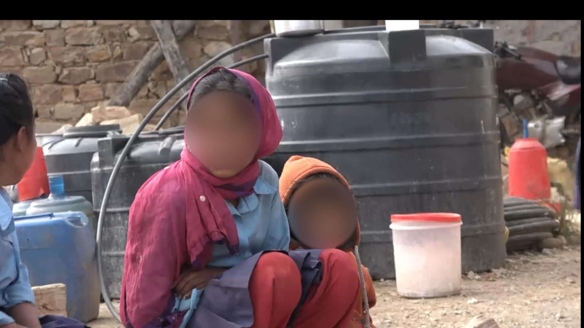 Zwei Kinder sitzen draußen auf dem Boden in der Nähe großer schwarzer Wassertanks. Ein Kind trägt einen rosa Schal und ein blaues Hemd, das andere eine orangefarbene Mütze. Ihre Gesichter sind unscharf. In der Nähe stehen ein weißer Eimer und weitere Gegenstände.