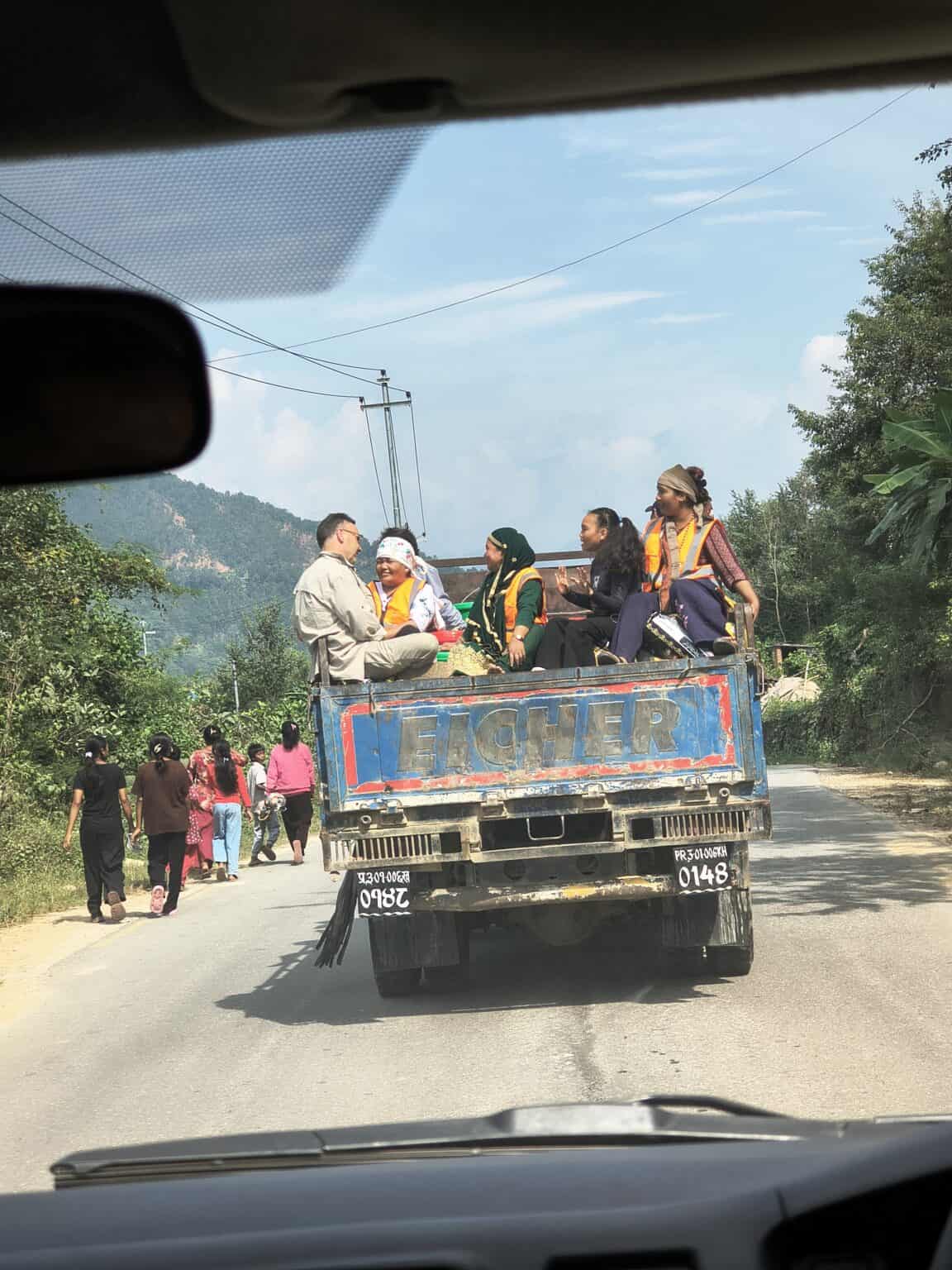 Eine Gruppe von Menschen sitzt auf der Ladefläche eines blauen Pick-ups, der eine Straße entlangfährt, während andere Menschen am Straßenrand entlanggehen.
