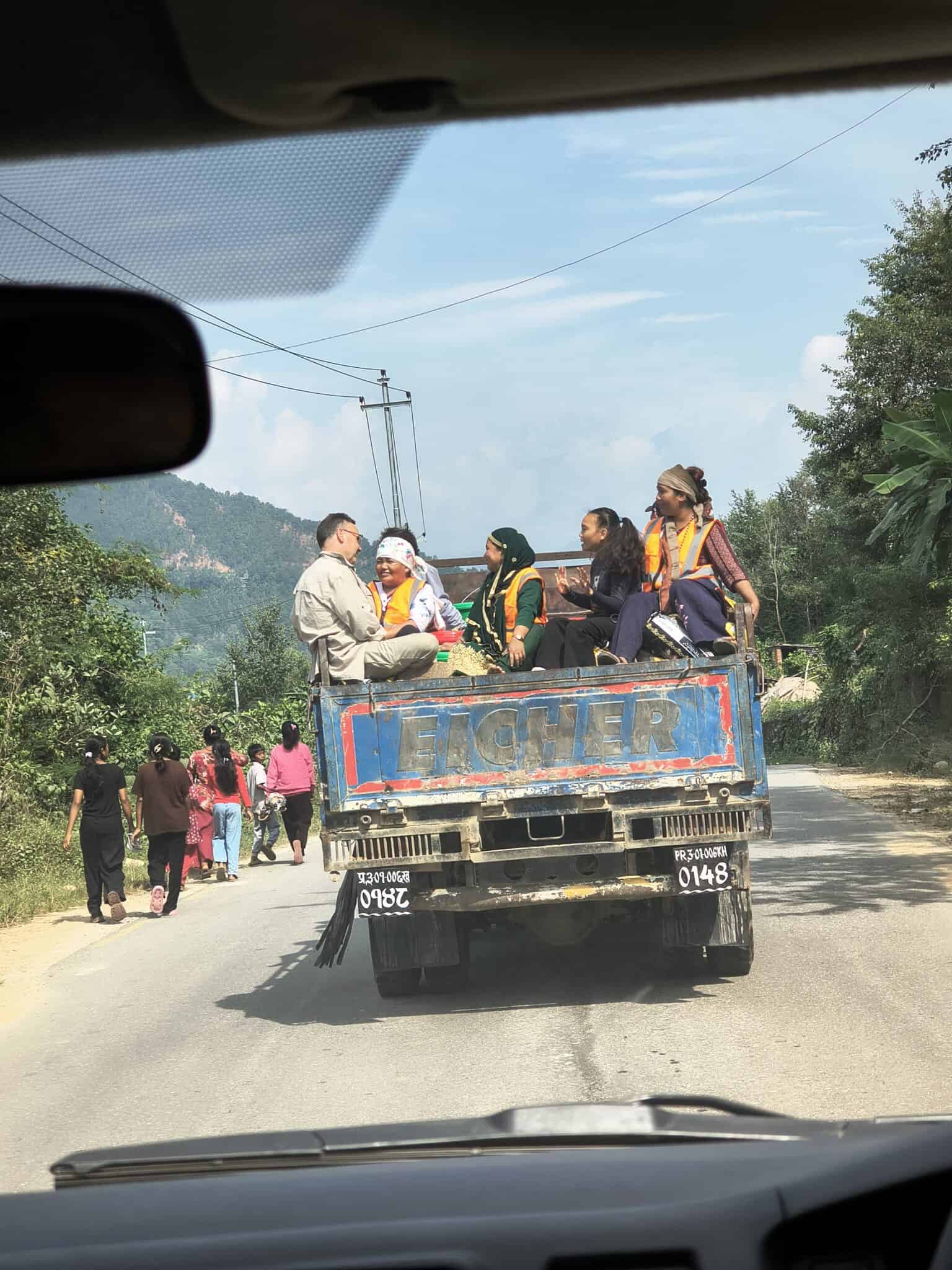 Eine Gruppe von Menschen sitzt auf der Ladefläche eines blauen Pick-ups, der eine Straße entlangfährt, während andere Menschen am Straßenrand entlanggehen.