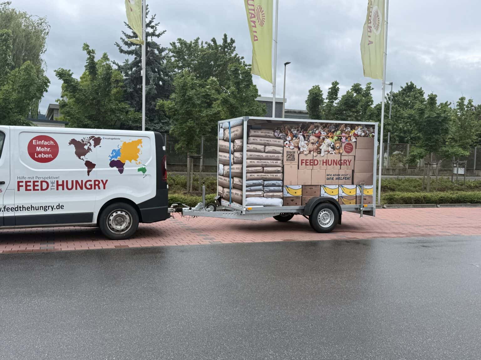 Ein Lieferwagen der Organisation „Feed the Hungry“, der einen Anhänger zieht, der mit Bildern von gestapelten Hilfsgütern wie Mehlsäcken, Lebensmittelkartons und Spielzeug beklebt ist.