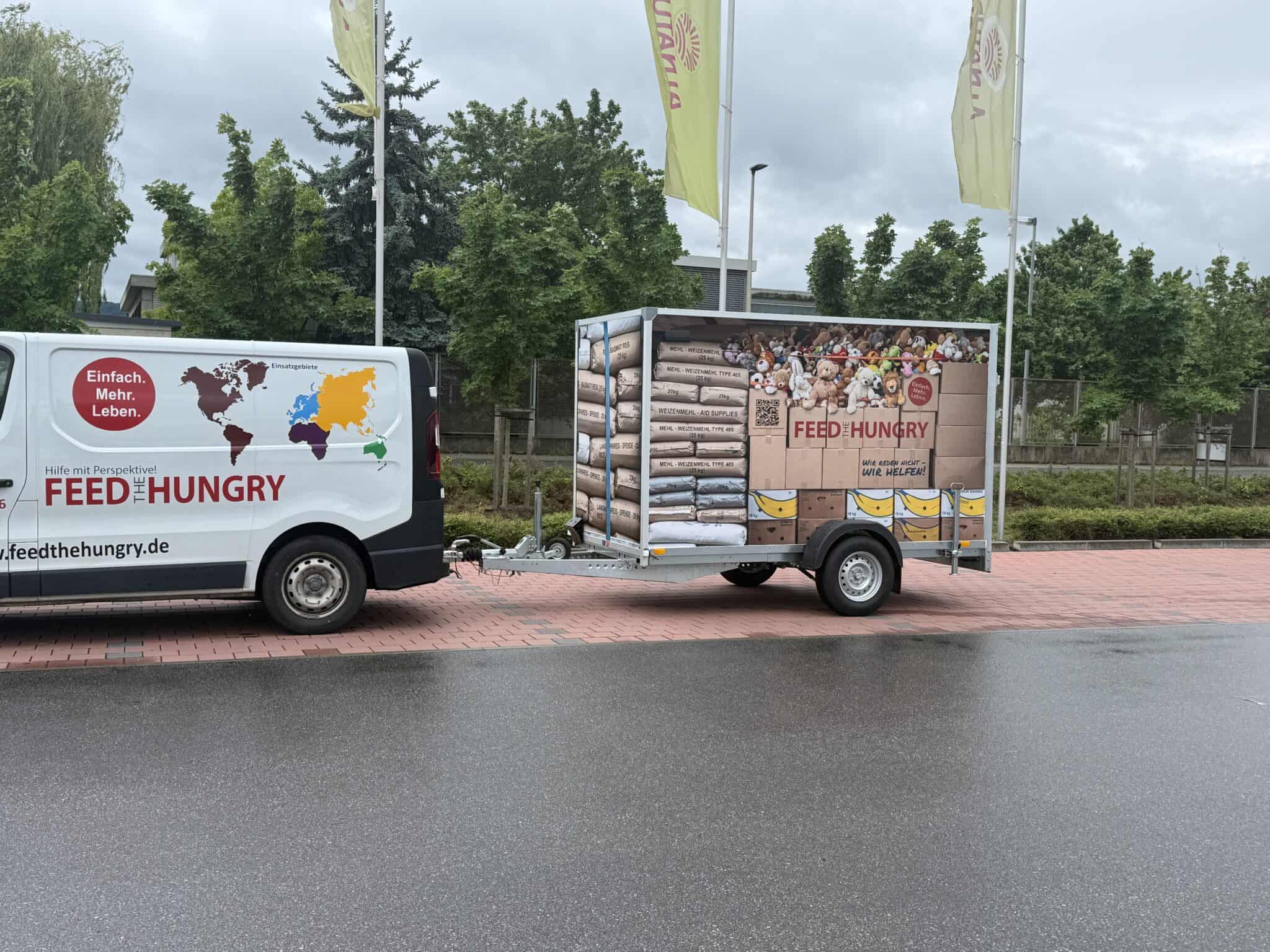 Ein Lieferwagen der Organisation „Feed the Hungry“, der einen Anhänger zieht, der mit Bildern von gestapelten Hilfsgütern wie Mehlsäcken, Lebensmittelkartons und Spielzeug beklebt ist.